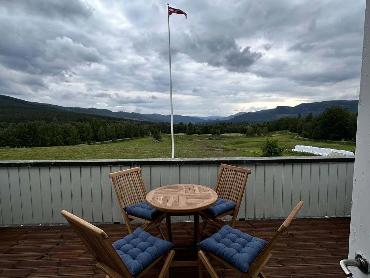 Verandaen på hovedhuset en sommerdag: teakbord og fire stoler, flaggstanga rett foran, og bak den beitet, dalen og fjellene